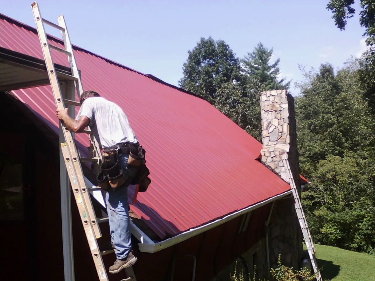 Roofer on a ladder beside a red metal roof and stone chimney — Soffit & Fascia Repair by Roofing Brothers in Alexandria